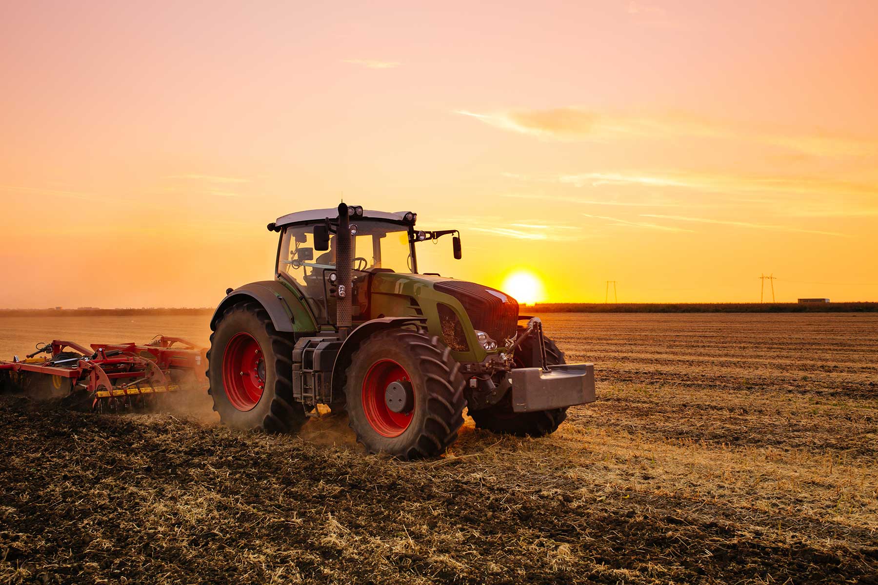 A green tractor stands in a field with the sun setting behind it, casting a warm golden glow across the landscape and creating long shadows on the grass
