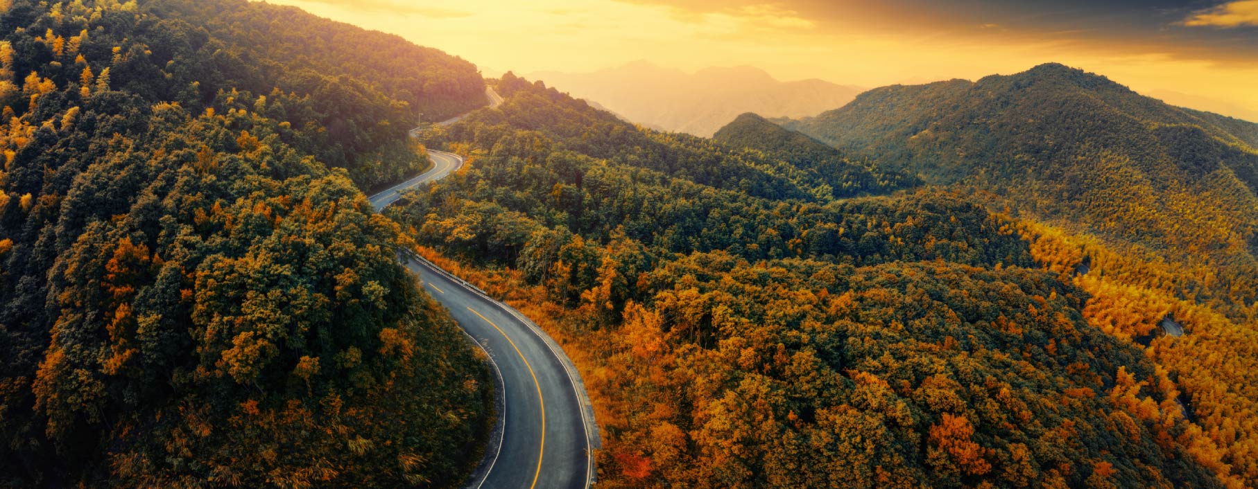 A scenic road winds through the Colorado mountains, surrounded by vibrant fall foliage in shades of red, orange, and gold, with rugged peaks rising in the background under a clear sky