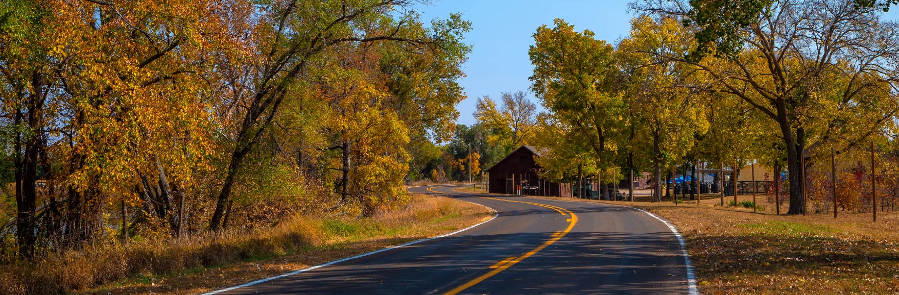 A winding country road in Nebraska curves through a landscape filled with autumn leaves, with trees in shades of orange, yellow, and red lining both sides under a crisp, clear sky.
