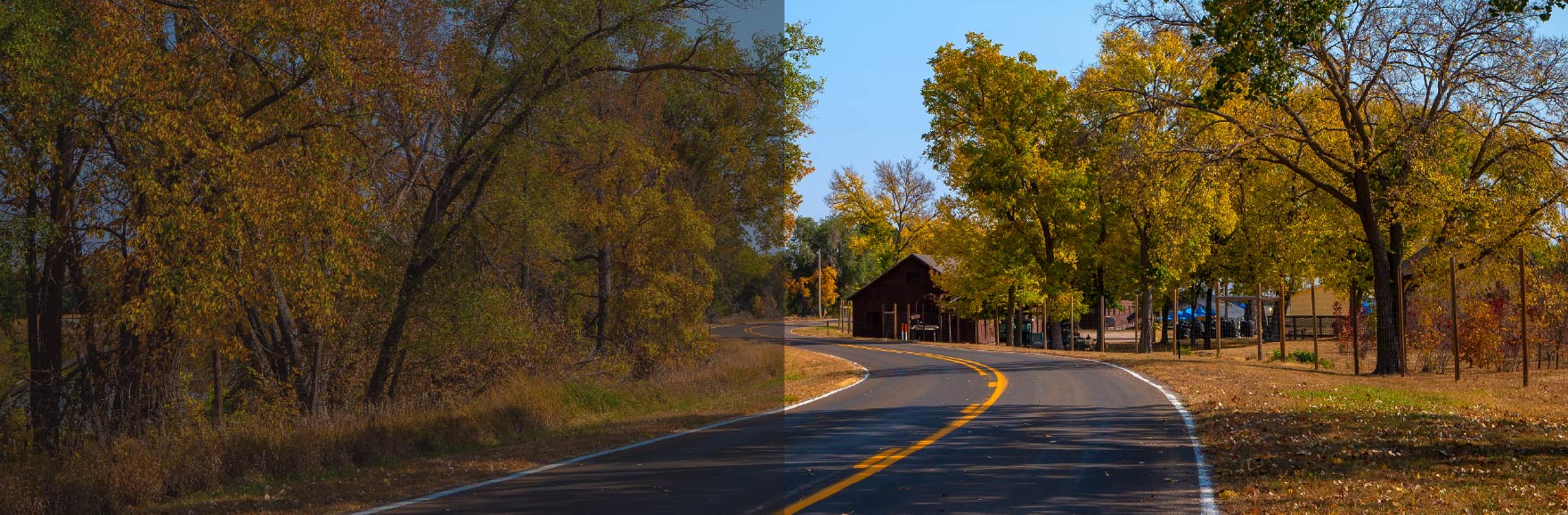 A winding country road in Nebraska curves through a landscape filled with autumn leaves, with trees in shades of orange, yellow, and red lining both sides under a crisp, clear sky.